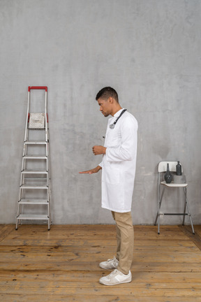 Side view of a young doctor standing in a room with ladder and chair showing a size of something