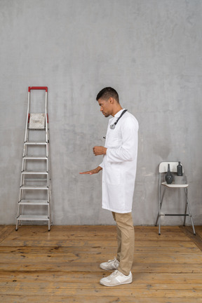 Vista lateral de un joven médico de pie en una habitación con escalera y silla mostrando el tamaño de algo