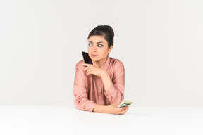 Молодая индийская женщина держит деньги счета и разговаривает по телефону
