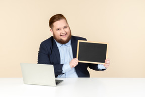 Lachender junger mann, der am schreibtisch sitzt und eine tafel hält