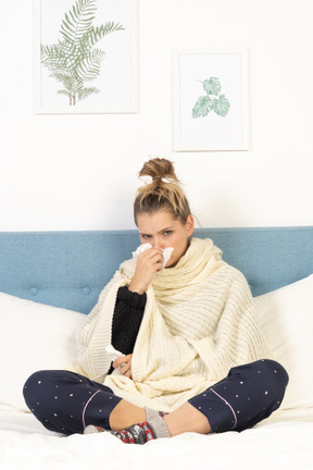 Front view of a young lady wrapped in white blanket sitting in bed and blowing nose
