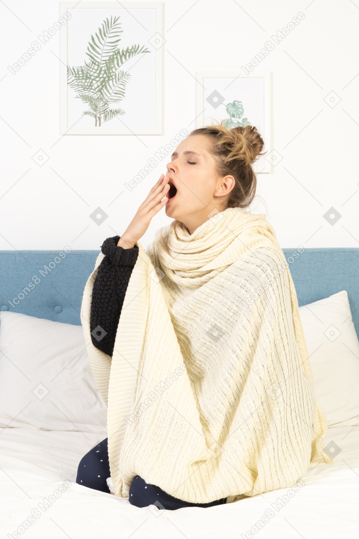Three-quarter view of a yawning young lady wrapped in white blanket sitting in bed