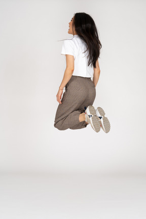 Vista posteriore di tre quarti di una giovane donna che salta in calzoni e t-shirt piegando le ginocchia