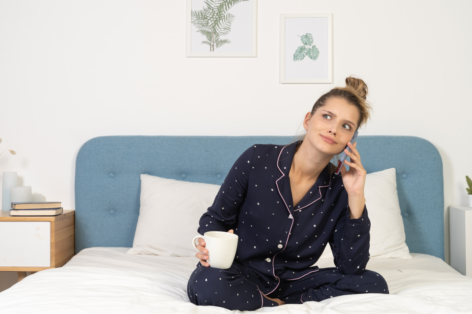 Front of a young perplexed female in pajama sitting in bed holding the cup and having a phone call