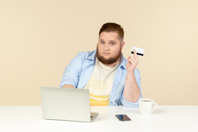 Jovem homem com excesso de peso, sentado no laptop e segurando o cartão do banco