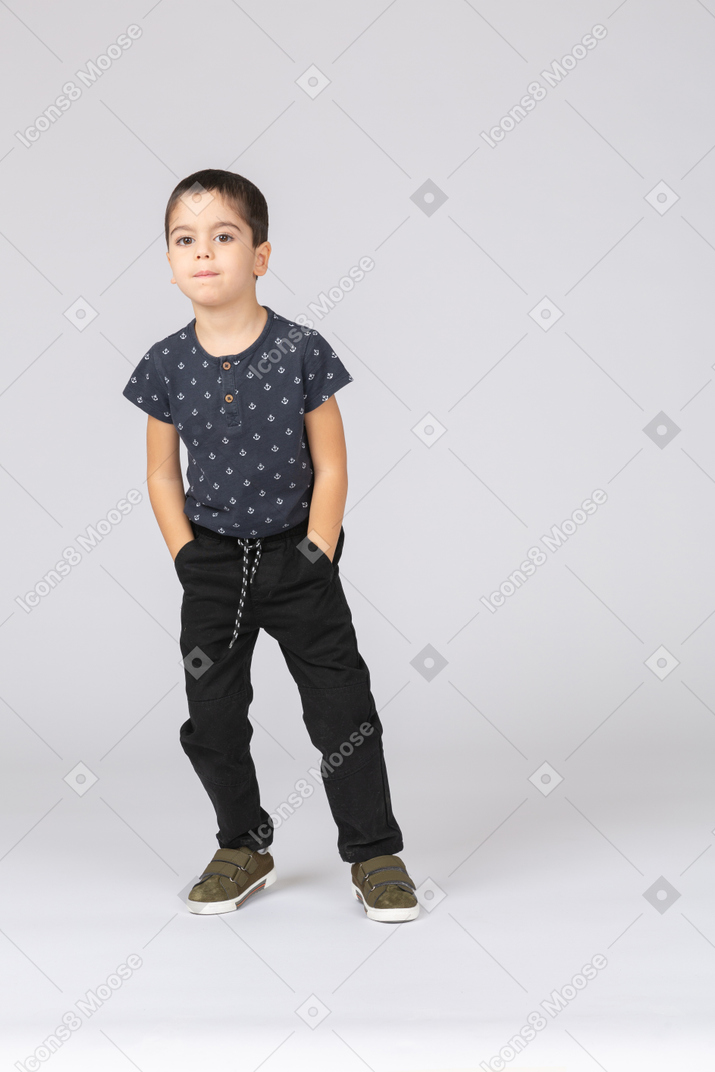 Vista frontal de um lindo menino em roupas casuais, posando com as mãos nos bolsos e olhando para a câmera