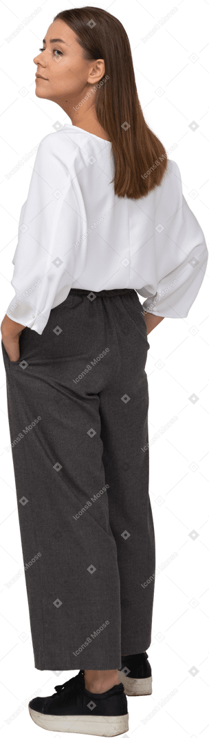 Vista posterior de tres cuartos de una joven en ropa de oficina poniendo las manos en los bolsillos y mirando a un lado