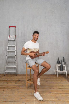 Three-quarter view of a man on a stool playing ukulele and smiling