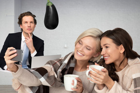 Two women taking a selfie while drinking coffee and a man looking at an eggplant