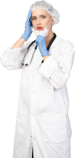 Vue de trois quarts d'une jeune femme médecin mettant un masque et regardant la caméra