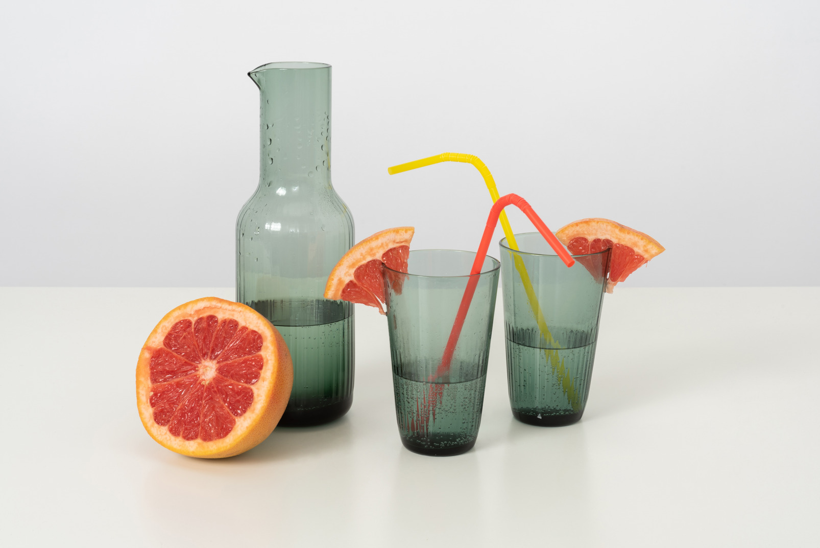 A glass bottle and glasses with some cold water and slices of a grapefruit, along with some colourful plastic straws