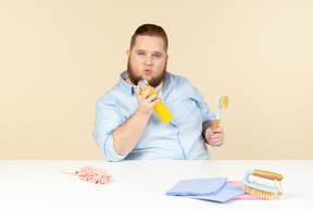 Zufriedener junger übergewichtiger mann, der am tisch sitzt und reinigungsgeräte hält