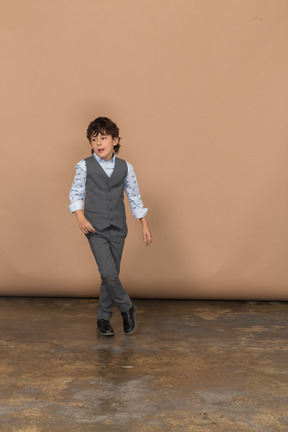 Vista frontal de un niño con traje gris de pie con las piernas cruzadas