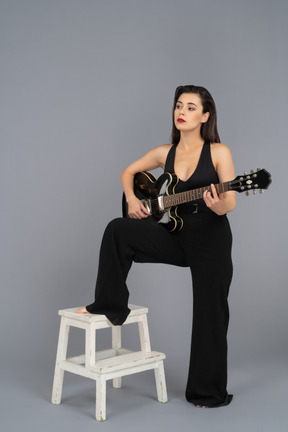 Bella giovane donna che suona una chitarra nera mantenendo la gamba su uno sgabello