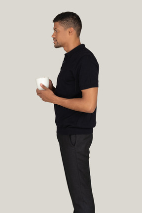 Standing in profile young man in black t-shirt and pants drinking coffee