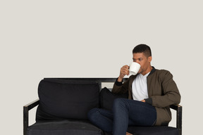 Front view of a young dreaming man sitting on a sofa while drinking coffee