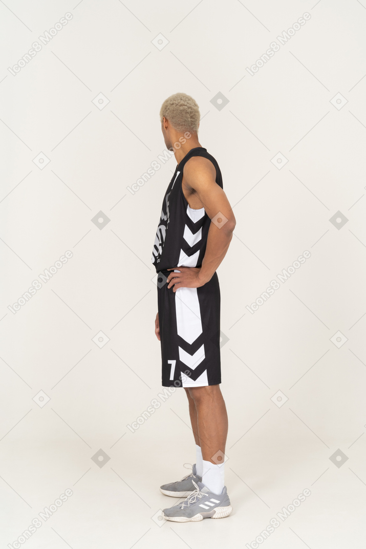 Vista lateral de un joven jugador de baloncesto masculino poniendo las manos en las caderas y girando la cabeza