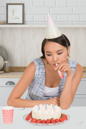 Sad woman in party hat blowing a party horn near birthday cake