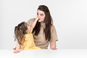 Kid girl sitting at the table and holding his mother nose