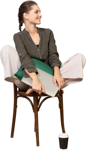 Front view of a smiling young woman sitting on a chair and holding her laptop