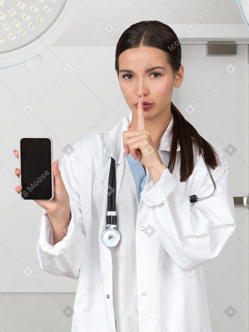 A female doctor holding a smartphone and making a quiet hand gesture