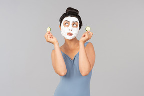 Woman with white facial mask holding cucumber slices