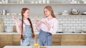 A teenage girl holding a glass with lemonade and standing next to a girl in a kitchen