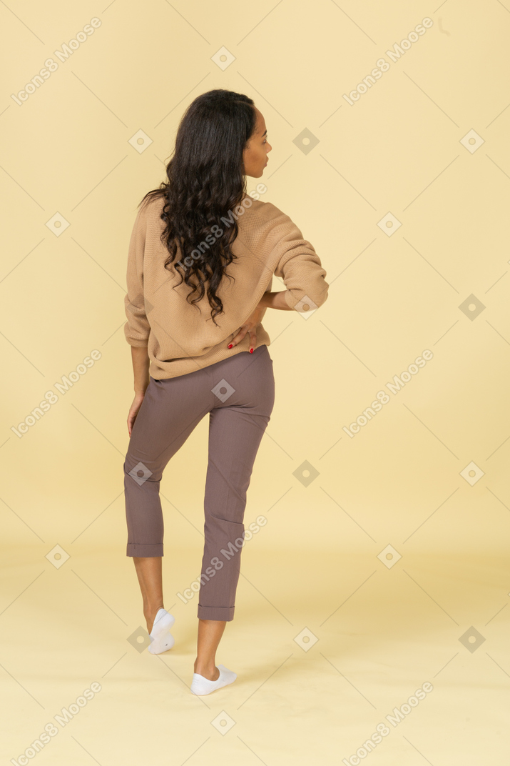 脇を見ながら腰に手を置く浅黒い肌の若い女性の背面図