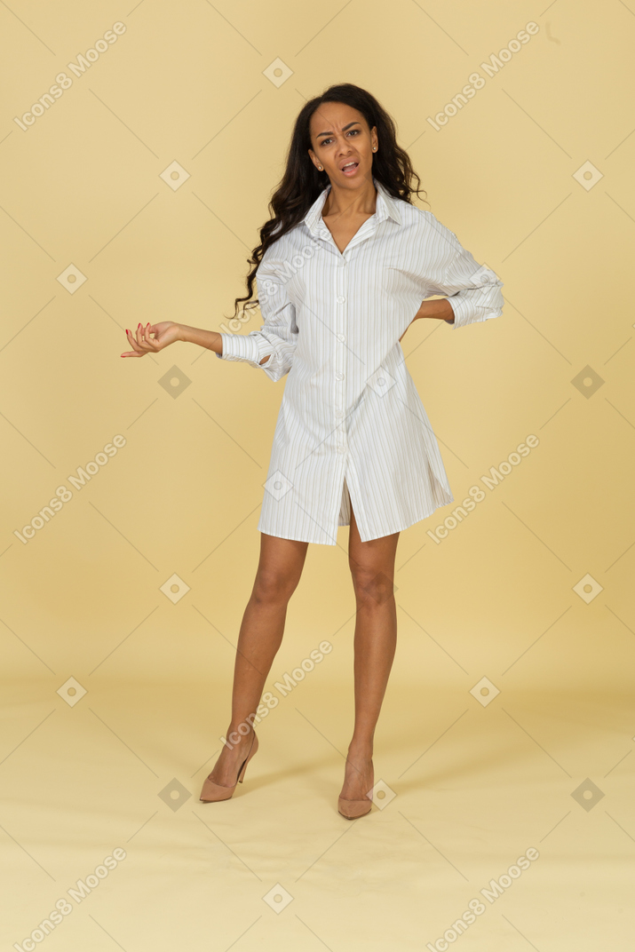 Vista frontal de una mujer joven de piel oscura cuestionando en vestido blanco poniendo la mano en la cadera