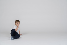 A young boy sitting on the ground with his hand on his mouth