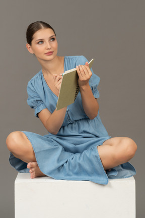 Front view of young woman sitting on a cube with marker and notebook