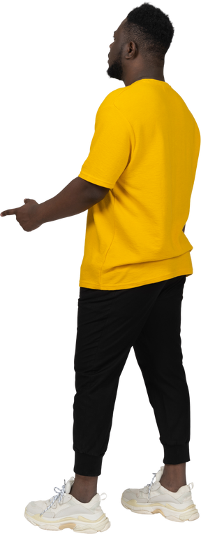 Vista posteriore di tre quarti di un giovane uomo dalla pelle scura con una maglietta gialla che indica il dito