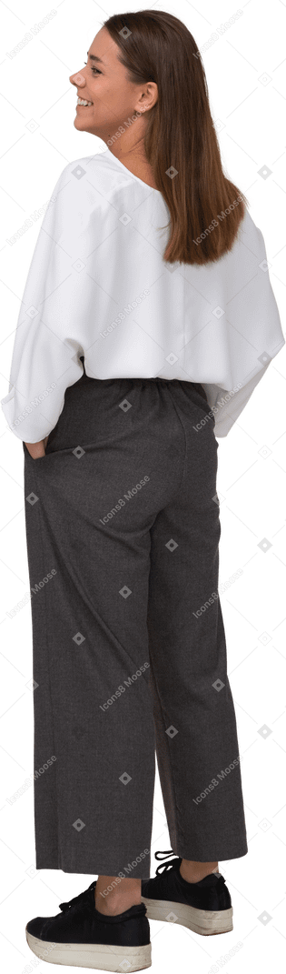 Vista posterior de tres cuartos de una joven sonriente en ropa de oficina poniendo las manos en los bolsillos