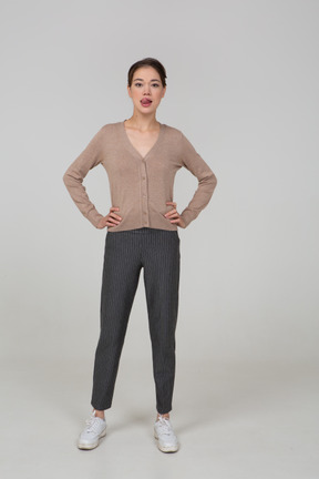 Vista frontale di una giovane donna in pullover e pantaloni che mette le mani sui fianchi e mostra la lingua