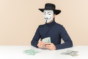 Hacker wearing vendetta mask sitting at the table and counting money