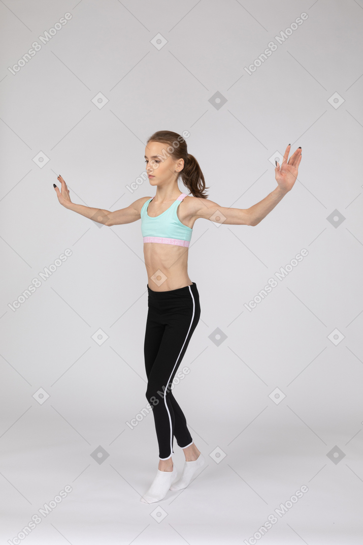 Three-quarter view of a teen girl in sportswear balancing on tiptoes while raising hands