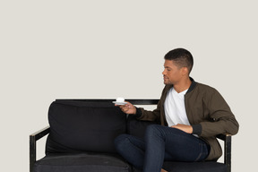 Three-quarter view of a young dreaming man sitting on a sofa with a cup of coffee