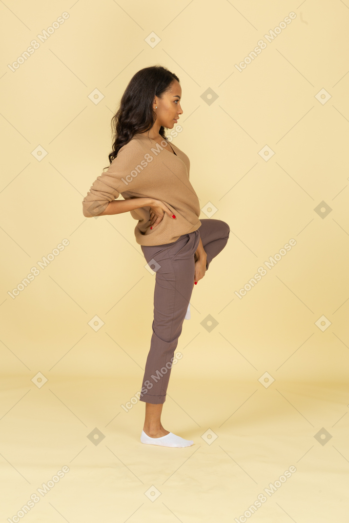 Vista lateral de uma jovem mulher de pele escura colocando a mão no quadril enquanto levanta a perna