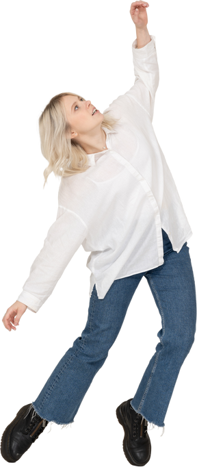 Front view of a blonde female in casual clothes dancing on her tiptoes and raising hand while looking up