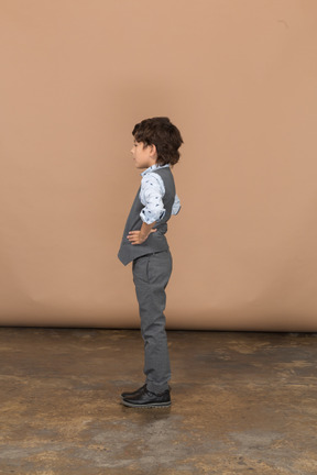 Вид сбоку на симпатичного мальчика в костюме, стоящего с руками на бедрах