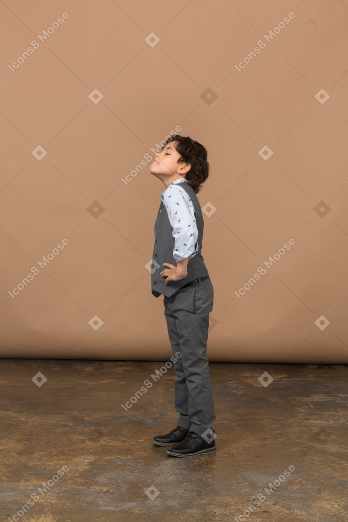 Vista lateral de um menino bonito de terno cinza em pé com as mãos nos quadris e olhando para cima