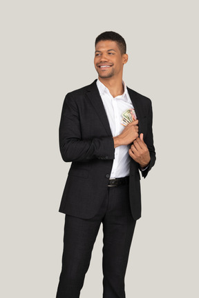 Вид спереди молодого человека в черном костюме, кладущего банкноты в карман