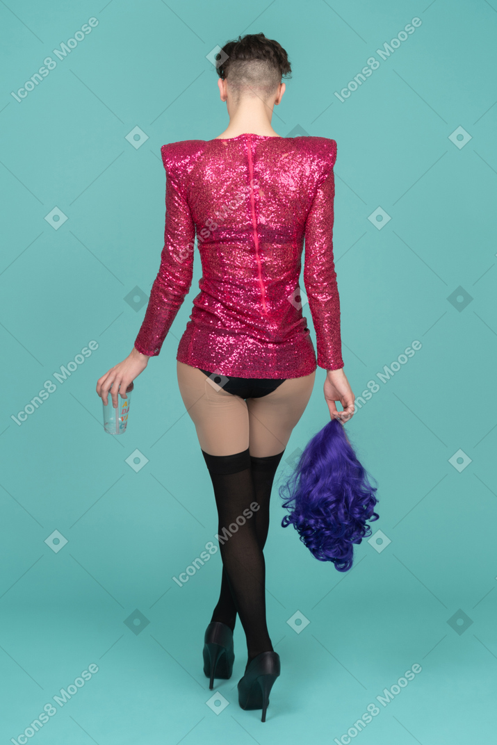 Vista trasera de una drag queen con un vestido rosa de lentejuelas sosteniendo una peluca y un vaso de plástico