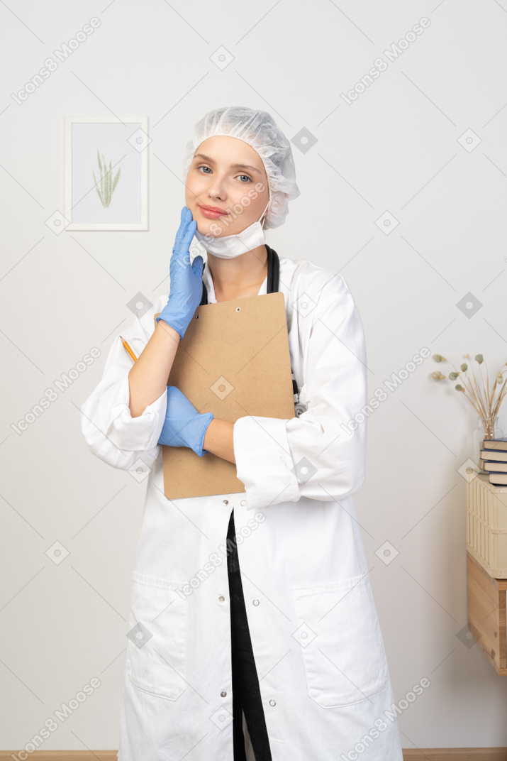 Front view of a pleased young female doctor holding pencil and tablet & touching face