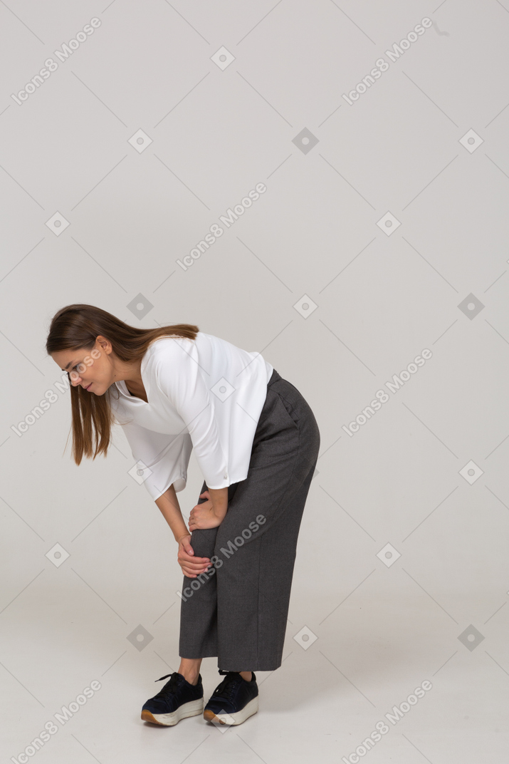 Three-quarter view of a young lady in office clothing bending down & touching knee