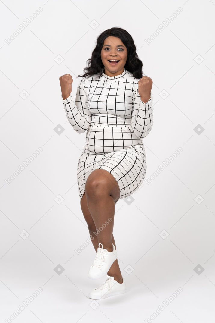 Vue de face d'une jeune femme heureuse dans des vêtements décontractés debout sur une jambe avec les mains vers le haut