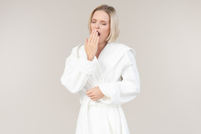 Young woman in bathrobe yawning and closing mouth with hand