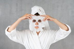 Mujer en bata de baño sosteniendo una máscara de hoja sobre la cara