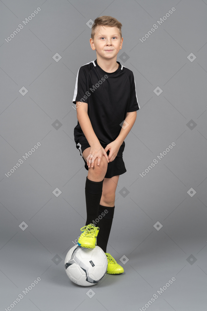 Vista frontale di un ragazzo bambino sorridente in uniforme di calcio mettendo il piede sulla palla e toccando il ginocchio