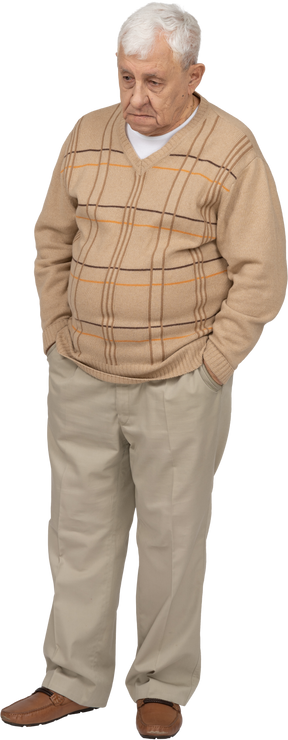 Vista frontal de un anciano con ropa informal parado con las manos en los bolsillos y mirando algo con interés